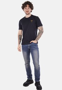 T-shirt di cotone blu navy con collo rotondo, maniche corte e un piccolo design ricamato sul petto, abbinata a jeans blu chiaro e sneakers nere.