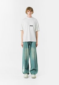 Camiseta blanca de manga corta con un bolsillo en el pecho y un gráfico de "MWM", combinada con pantalones de denim azules claros de corte holgado que presentan letras en los lados.