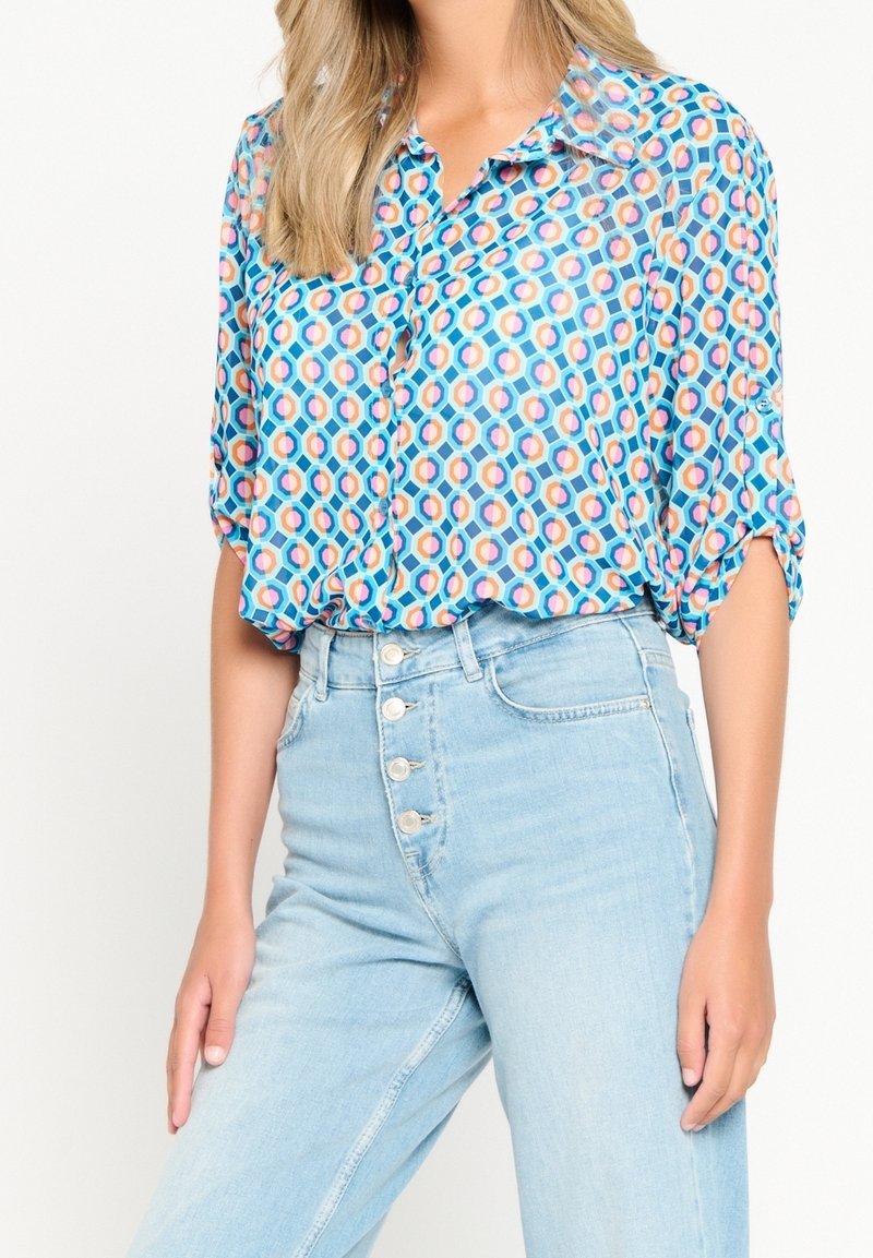 Camicia a maniche corte con bottoni di colore blu, decorata con un motivo geometrico circolare in rosa e arancione. Indossata con jeans chiari a vita alta.