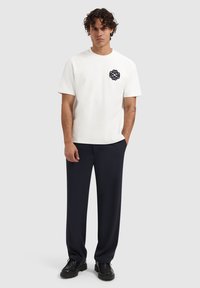 Witte katoenen t-shirt met een marineblauwe geborduurde design op de linkerborst, gecombineerd met donkerblauwe broek en zwarte lace-up schoenen.