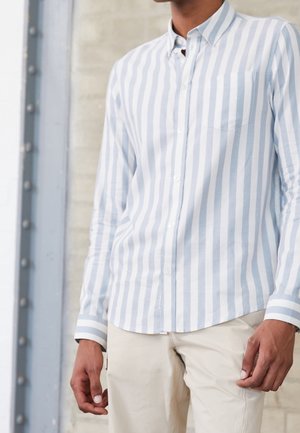 Homme portant une chemise à rayures verticales bleu clair et blanc à boutons et un pantalon beige, debout à l'intérieur contre un mur texturé.
