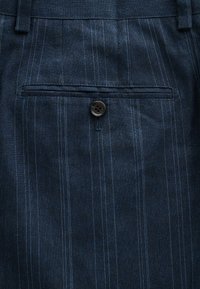 Tissu bleu marine à fines rayures avec une poche horizontale fendue sécurisée par un seul bouton noir et une boutonnière en dessous de la poche.