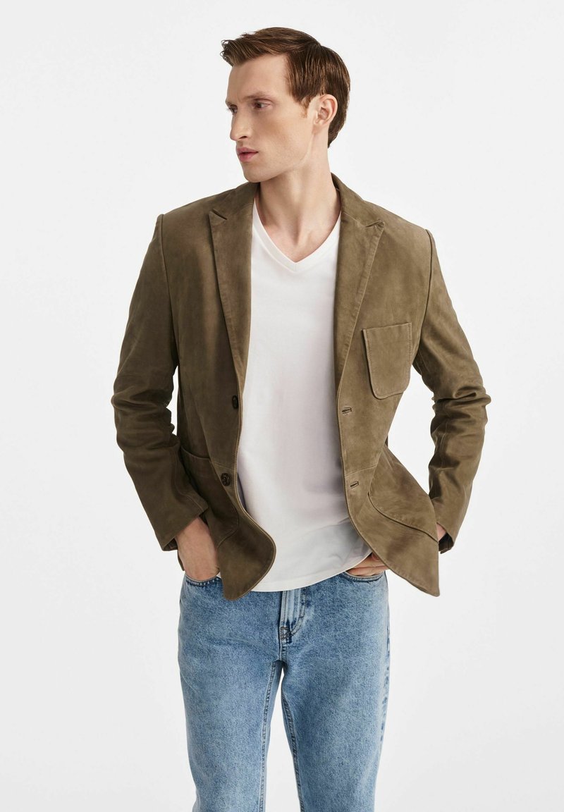 Khaki zamszowa marynarka z pojedynczą kieszenią na klatce piersiowej, dwoma kieszeniami z przodu i klapami, noszona na białej koszulce V-neck i niebieskich dżinsach.