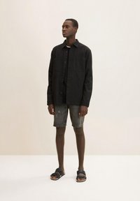Schwarzes, knopfbares Hemd über einem schwarzen T-Shirt, kombiniert mit verwaschenen grauen Shorts und dunklen Sandalen. Einfaches, lässiges Design.