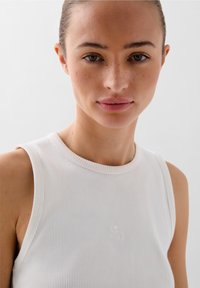 Bílý žebrovaný tílko s kulatým výstřihem a bezrukávovým designem, s jemně vyšitým logem v oblasti hrudníku.