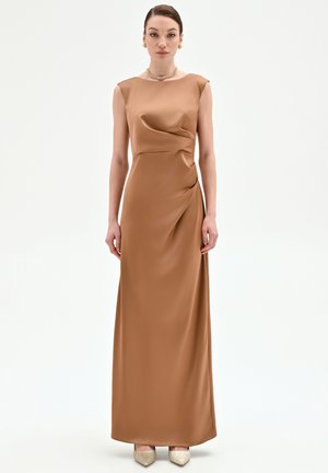 Donna in piedi che indossa un abito marrone in raso senza maniche fino a terra con dettagli arricciati, orecchini dorati, collana e scarpe metallizzate con punte appuntite.