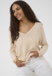 Pull beige à col en V avec manches longues, texture côtelée et coupe décontractée, associé à un pantalon blanc et un bracelet manchette doré.