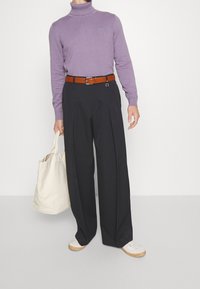 Person, der ein lavendelfarbenes Rollkragenoberteil, schwarze Schlaghosen mit einem braunen Gürtel, weiße Sneakers und eine beige Tragetasche trägt.