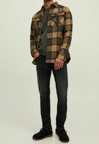 Uomo che indossa una camicia a quadri beige e nera sopra un maglione grigio, jeans neri e scarpe nere, in piedi con una mano in tasca.