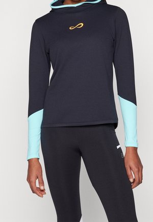 Femme portant un sweat à capuche noir à manches longues avec des poignets de manches bleu clair et un symbole infini orange sur la poitrine, associée à des leggings noirs.