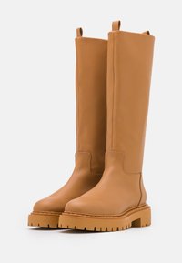 Botas de cuero color marrón claro hasta la rodilla, con un acabado suave, suela gruesa y texturizada, y adornos en forma de lazo en la parte superior.