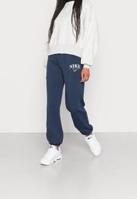 Grå oversized sweatshirt, marinblå träningsoverallsbyxor med vit "NIKE" logo, och vita sneakers med luftkuddar. Avslappnad passform, tapered cuffs.