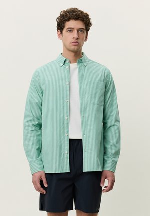Man with curly hair wearing a light green striped button-up shirt over a white t-shirt and black shorts against a plain background.