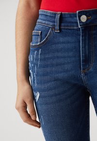 Μπλε τζιν παντελόνι ψηλής μέσης με φθαρμένα σημεία, μπροστινή τσέπη και πορτοκαλί ραφές. Κοντινή λήψη της ζώνης και του χεριού που ξεκουράζεται στο πόδι.