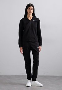 Zwarte zip-up hoodie en bijpassende broek van zachte stof. Met een minimalistisch logo op de borst. Genesteld met witte sneakers.