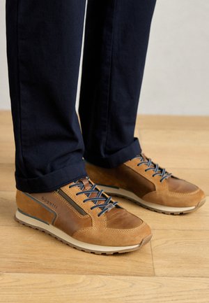 Brown and tan sneakers with a mix of smooth and textured leather, blue laces, and a light sole, worn with navy pants against a wooden floor.