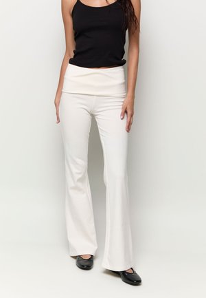 Femme portant un haut noir sans manches, un pantalon blanc évasé taille haute et des ballerines noires, debout devant un fond clair uni.