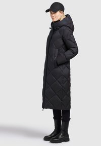 Long black quilted puffer coat with a hood, featuring a front zipper and side pockets, paired with black ankle boots and a matching cap.