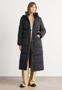 Γυναίκα που φοράει ένα μακρύ μαύρο παλτό με κούμπωμα, μπεζ πουλόβερ, φαρδιά μπεζ παν trousers και μαύρα παπούτσια, στέκεται με το ένα χέρι στην τσέπη.