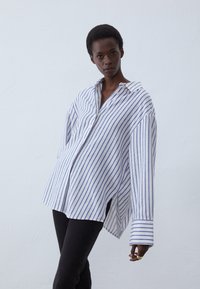 Striped button-up shirt in white and blue cotton with a relaxed fit, drop shoulders, and side slits, paired with black pants.
