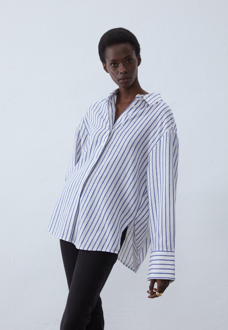 Striped button-up shirt in white and blue cotton with a relaxed fit, drop shoulders, and side slits, paired with black pants.