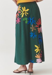 Fustă maxi florală verde cu modele de flori strălucitoare, multicolore. Material moale cu un elastic la talie. Încălțăminte: sandale maro cu bretele.