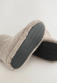 Beige, plüschige Hausschuhe mit einer strukturierten, flauschigen Außenseite und einer schwarzen Gummisohle mit Wellenmuster, entworfen für Komfort und Wärme.
