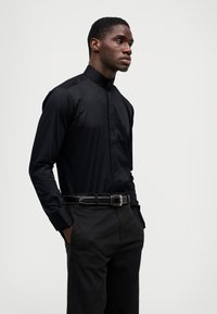 Camisa preta de manga longa com botões e colarinho alto, combinada com calças pretas e um cinto a condizer com detalhes de costura branca.