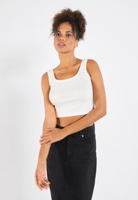 Hvit kort tanktopp med ribbestruktur, firkantet utringning og bølget kant. Kombinert med et høyhals svart denimskjørt.