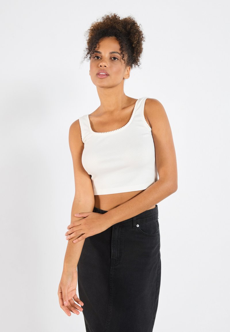 Hvit kort tanktopp med ribbestruktur, firkantet utringning og bølget kant. Kombinert med et høyhals svart denimskjørt.
