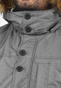 Homme portant un manteau d'hiver gris avec quatre boutons noirs et une capuche bordée de fourrure visible en haut du cadre.