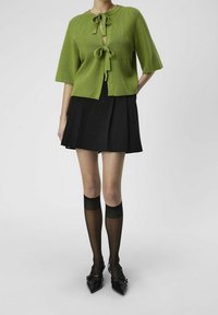 Cardigan en tricot vert à manches courtes, avec une fermeture à nouer au niveau du cou. Associé à une jupe noire plissée et des chaussettes noires transparentes jusqu'aux genoux.