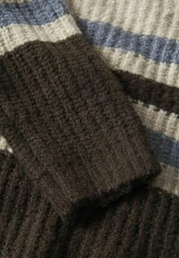 Nahaufnahme von gerippetem Strickstoff mit braunem Bündchen über gestreifter beiger und blauer Wollstruktur.