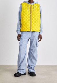 Persona che indossa un gilet trapuntato giallo brillante sopra una felpa grigia chiara e pantaloni cargo grigi chiari, con scarpe chunky nere, in piedi su un pavimento semplice.