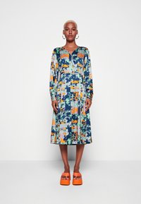 Vestido longo de mangas compridas colorido com um padrão floral em azul, verde, laranja e roxo, com decote em V e saia rodada; combinado com sapatos de salto laranja.