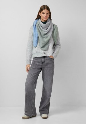Giovane donna indossa un maglione a maglia grigio, sciarpa multicolore blu e verde, jeans grigi larghi e sneakers beige, in piedi contro uno sfondo neutro.