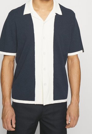 Homme portant une chemise à manches courtes boutonnée avec des blocs de couleur verticaux noirs et blancs et un col blanc.