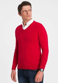 Czerwony sweter z dzianiny z dekoltem w serek i długimi rękawami, z delikatnym logo. Nosi się go na białej koszuli z kołnierzykiem i w połączeniu z niebieskimi dżinsami.