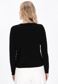 Vrouw met blonde golvende haren, gekleed in een zwart kabelgebreid sweater en witte broek, staand met de rug naar de camera tegen een effen achtergrond.