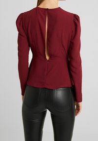 Blusa lunga borgogna con dettaglio delle spalle a palloncino, caratterizzata da una scollatura a chiave sul retro e chiusura con bottone. Indossata con pantaloni in pelle neri aderenti.