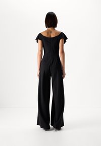 Melnā kombinezons ar īsām volānu piedurknēm, augstu kakla izgriezumu aizmugurē, platu kāju dizainu un slēptu ritentiņu aizmugurē.
