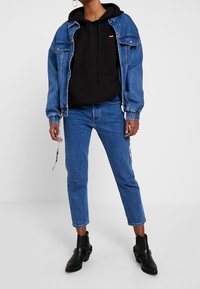 Chaqueta de mezclilla en azul claro sobre una sudadera negra, combinada con jeans cortos azules y botas negras de tobillo con hebillas.