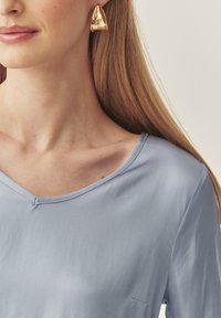 Helles blaues V-Ausschnitt-Blusenkleid mit einer glatten Textur. Verfügt über ein einfaches Design und kurze Ärmel. Goldene dreieckige Ohrringe vervollständigen den Look.