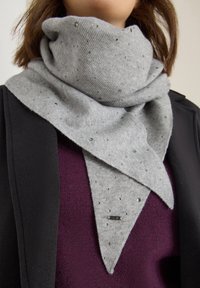 Sciarpa grigia con una texture a coste, adornata con piccoli accenti riflettenti, sagomata in un design triangolare, indossata sopra un top viola.