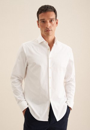 Homme avec des cheveux courts et foncés portant une chemise blanche à boutons et un pantalon sombre, se tenant les mains dans les poches devant un fond beige.
