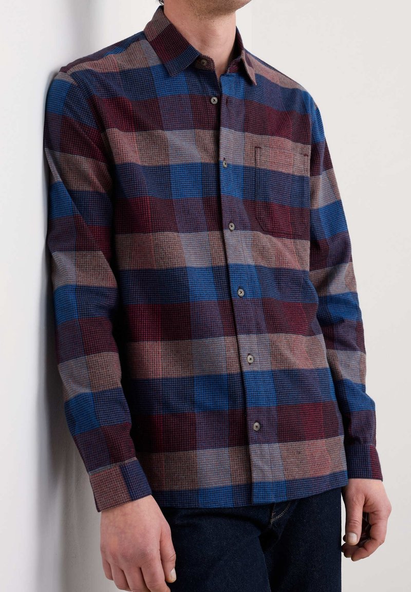 Homme portant une chemise à manches longues boutonnée avec un grand motif à carreaux bleu, bordeaux et gris, debout contre un mur blanc.