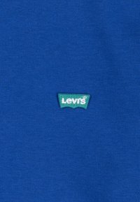 Kék pamut ing, amelyen egy kis zöld hímzett Levi's logó folt található a mellrészen. Az anyag sima, kötött textúrájú.