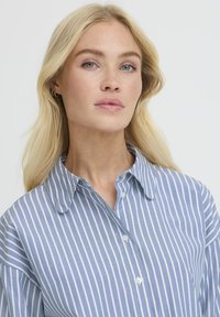 Blonde Frau mit blauen Augen, die ein blauen und weißen vertikal gestreiften Hemd trägt, sieht leicht neben die Kamera mit einem neutralen Ausdruck.