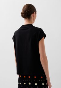 Femme avec une coiffure en chignon, portant un haut noir sans manches et une jupe noire à motifs floraux orange et blanc, debout devant un fond uni.