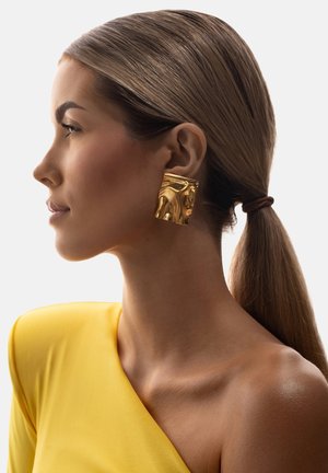 Kobieta z gładkim kucykiem, ubraną w duży, teksturowany, złoty kwadratowy kolczyk oraz żółtą bluzkę odsłaniającą ramiona na jednolitym tle.